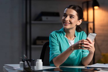Üniformalı gülümseyen hemşire masada oturuyor ve gece vardiyasında akıllı telefon kullanıyor.