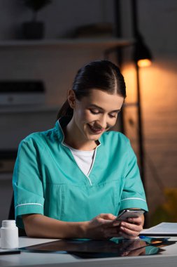 Gece vardiyasında üniformalı çekici bir hemşire masada oturuyor ve akıllı telefon kullanıyor.