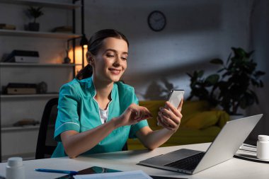 Üniformalı gülümseyen hemşire masada oturuyor ve gece vardiyasında akıllı telefon kullanıyor.