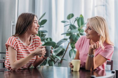 cheerful daughter and mother sitting at kitchen table, talking and drinking tea clipart
