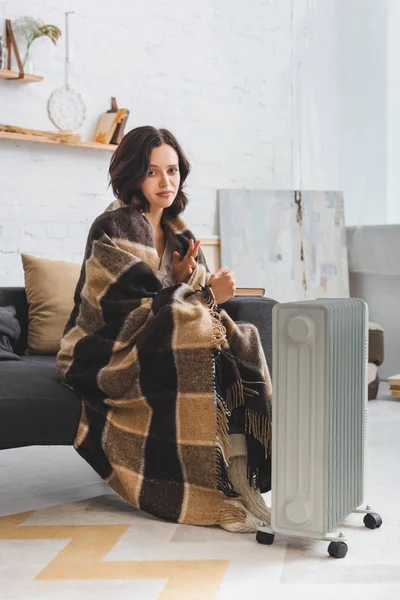 beautiful cold woman with blanket warming up near heater