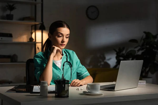 Ελκυστική Νοσοκόμα Στολή Κάθεται Στο Τραπέζι Και Κοιτάζοντας Φορητό Υπολογιστή — Φωτογραφία Αρχείου