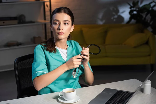 Enfermera Atractiva Uniforme Sentada Mesa Sosteniendo Estetoscopio Durante Turno Noche — Foto de Stock