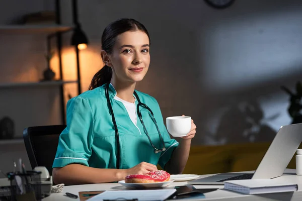 Χαμογελαστή Νοσοκόμα Στολή Κάθεται Στο Τραπέζι Και Κρατώντας Κύπελλο Κατά — Φωτογραφία Αρχείου