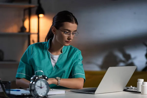Enfermeira Atraente Uniforme Óculos Sentados Mesa Olhando Para Laptop Durante — Fotografia de Stock