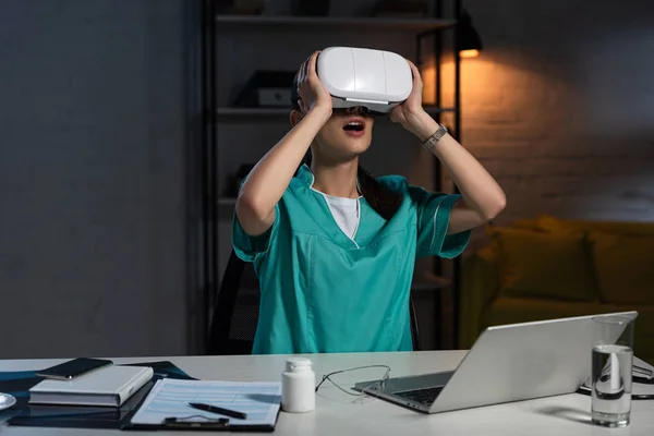 Шокированная Медсестра Форме Гарнитурой Виртуальной Реальности Сидящая Столом Время Ночной — стоковое фото