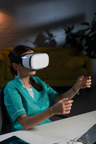 nurse in uniform sitting at table with virtual reality headset during night shift
