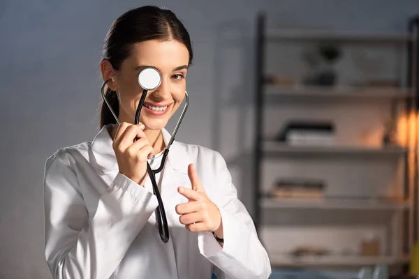 Médico Sorridente Casaco Branco Segurando Estetoscópio Apontando Com Dedo Durante — Fotografia de Stock