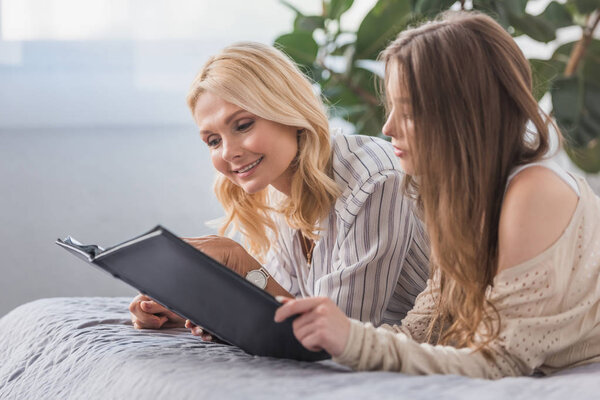 happy mother and daughter lying on bed and looking at photo album