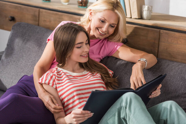 happy mother and daughter sitting on sofa and looking at photo album together