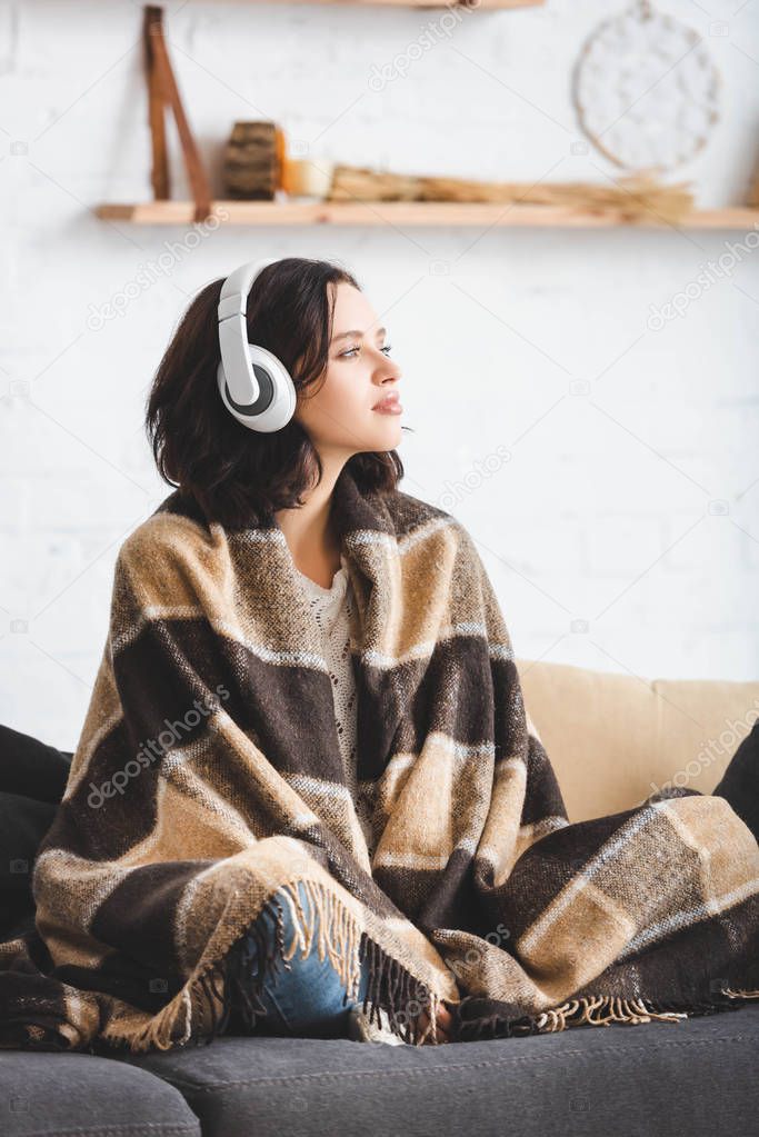 beautiful girl thinking and listening music with headphones while sitting in blanket 