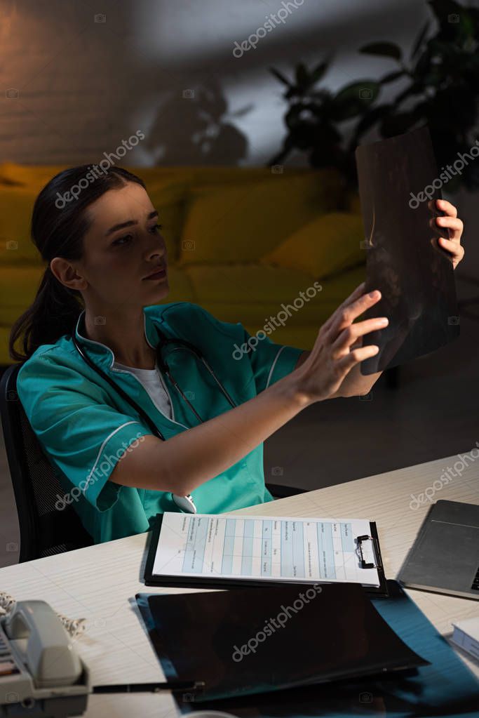 Attractive nurse in uniform sitting at table and looking at x-ray scan during night shift