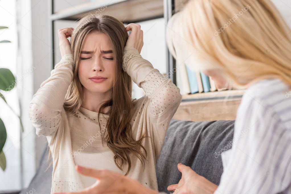 upset girl plugging ears with hands while sitting near talking mother