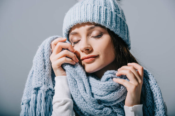 tender girl posing with closed eyes in blue knitted hat and scarf, isolated on grey