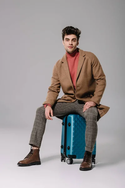 穿着米色外套的英俊男子坐在灰色行李箱上 — 图库照片