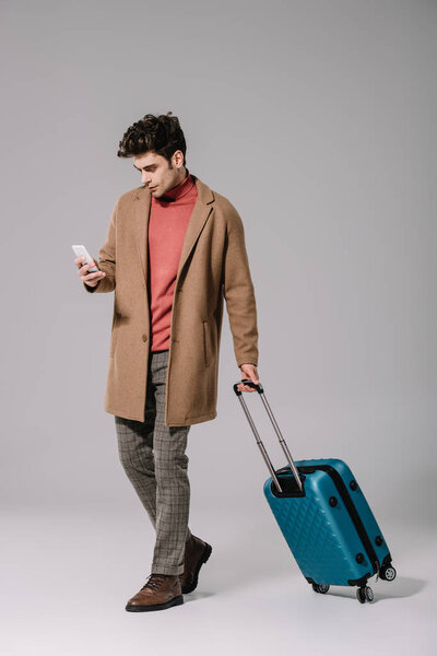 стильный мужчина в бежевом пальто с дорожной сумкой с помощью смартфона на сером

