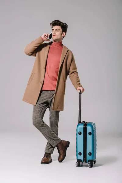 穿着米黄色外套的时髦男人 一边用智能手机说话 一边拿着灰色旅行袋 — 图库照片