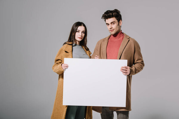 fashionable couple in beige coats posing with empty board, isolated on grey