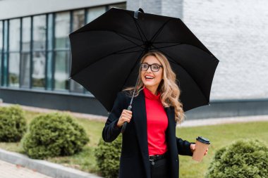 Siyah ceketli, elinde şemsiye ve kağıt bardak tutan çekici ve gülümseyen bir iş kadını. 
