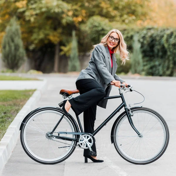 コートやメガネを着た魅力的なビジネスウーマンが自転車に乗ったり笑ったり — ストック写真