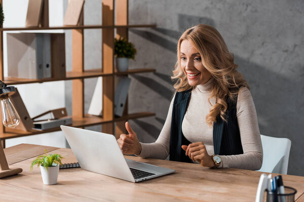 attractive and smiling businesswoman showing thumb up and looking at laptop 