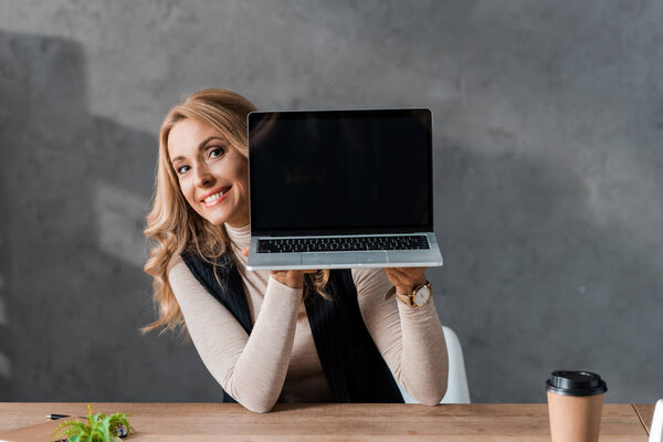 attractive and smiling businesswoman sitting at table and holding laptop 