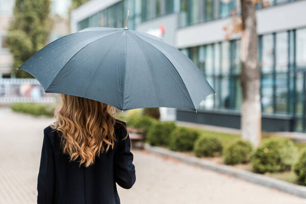 back view of blonde businesswoman in black coat holding umbrella 