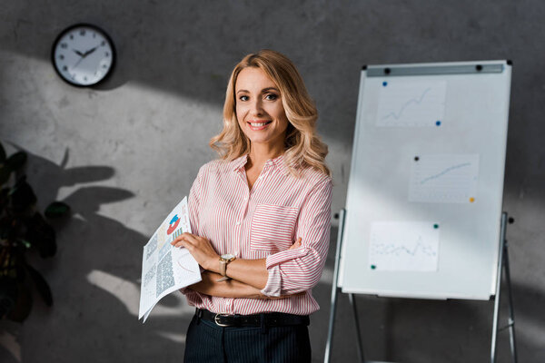 attractive and blonde businesswoman in shirt smiling and holding papers 