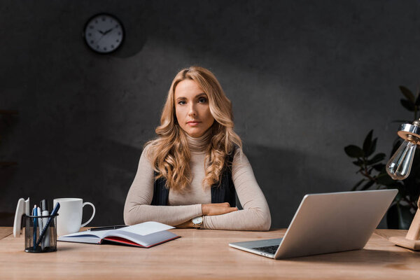 attractive and blonde businesswoman sitting at table and looking at camera in office 