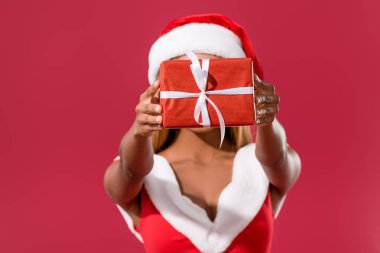 Noel Baba şapkası ve noel elbisesi giymiş genç Afro-Amerikalı kız kırmızı üzerine izole edilmiş bir şekilde yüzünün önünde hediye paketi tutuyor.