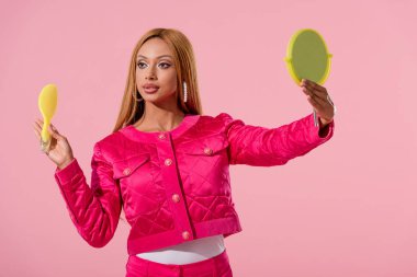 attractive, stylish african american woman holding hairbrush and mirror isolated on pink, fashion doll concept clipart
