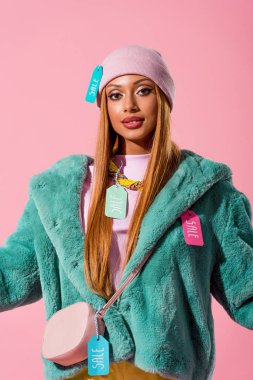 Giysilerinde indirimli etiketleri olan şık Afro-Amerikalı kadın pembe, moda bebeği konseptinde izole edilmiş kameraya bakıyor.