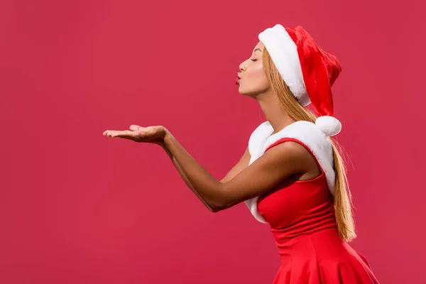 身着圣诞礼服头戴圣诞礼帽的非洲裔美国女孩的侧面照片 在红色的天空中发出隔离的空气之吻 — 图库照片