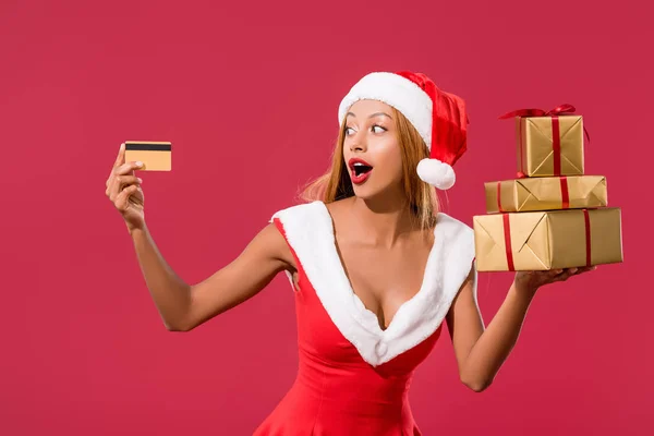 身穿圣塔帽和圣诞礼服的兴奋的非洲裔美国女孩 手持信用卡和礼品盒 与红色隔离 — 图库照片