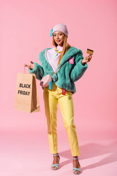 cheerful, stylish african ameican girl holding credit card and shopping bag with black friday inscription on pink background, fashion doll concept