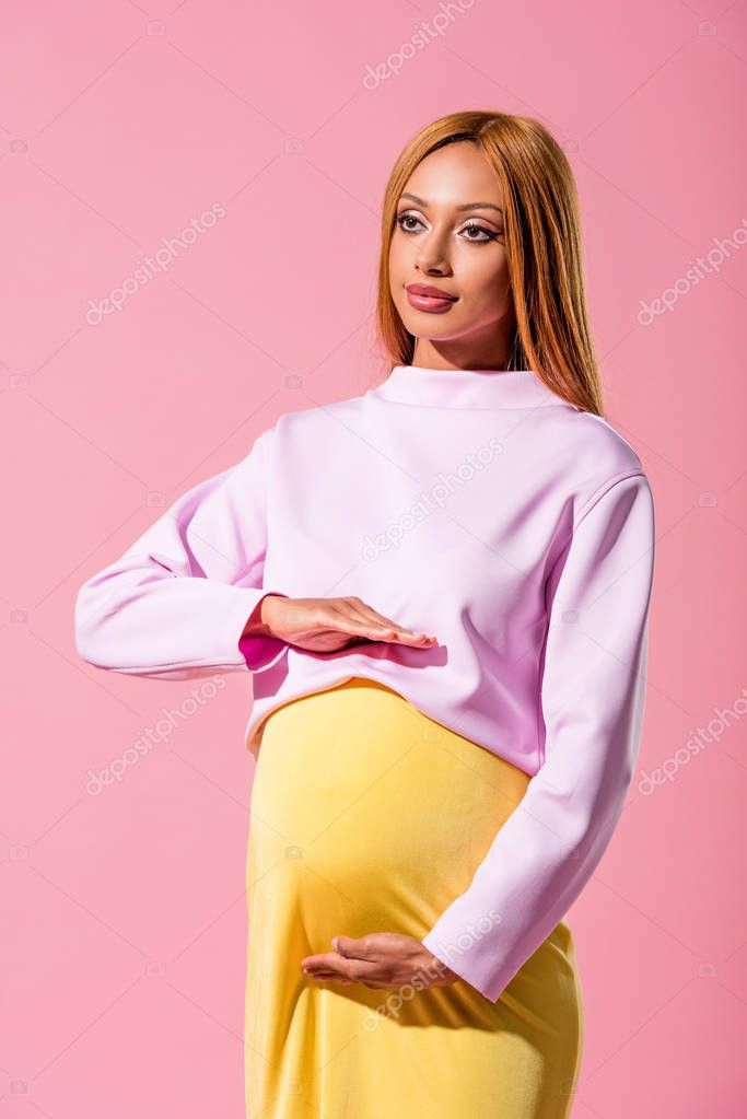 trendy, pregnant african american woman touching tummy and looking away isolated on pink, fashion doll concept