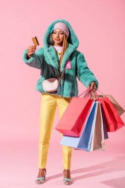 Pembe arka planda kredi kartı ve alışveriş torbaları tutan şık Afrikalı Amerikalı kadın, moda bebeği konsepti.