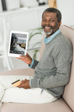 Gülümseyen Afrikalı Amerikalı adam kredi kartı tutuyor ve ekranda bilet başvurusu uygulaması ile dijital tablet gösteriyor.
