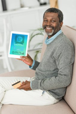 Gülümseyen Afrikalı Amerikalı adam kredi kartı tutuyor ve ekranda online alışveriş uygulaması ile dijital tablet gösteriyor