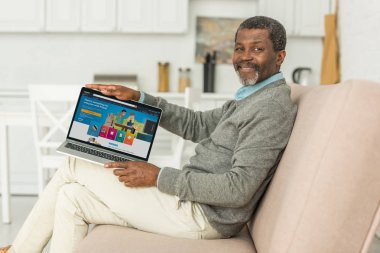 Kyiv, Ukrayna - 2 Ekim 2019: Afro-Amerikalı neşeli adam kanepede oturuyor ve ekranda Amazon uygulaması olan bir dizüstü bilgisayar gösteriyor.