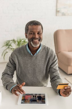 Neşeli Afrikalı Amerikalı adam kredi kartı tutuyor ve ekranda uygulamalı dijital tablet kullanıyor.