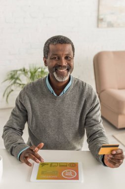 Neşeli Afrikalı Amerikalı adam kredi kartı tutuyor ve ekranda online alışveriş uygulaması olan dijital tablet kullanıyor.