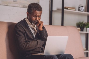 Üzgün Afrikalı Amerikalı bir adam kanepede otururken dizüstü bilgisayara bakıyor.