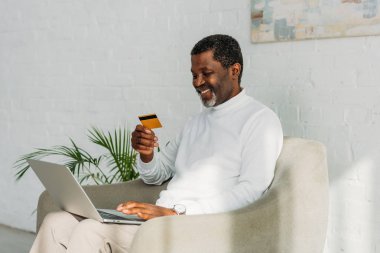 Neşeli Afrikalı Amerikalı adam dizüstü bilgisayar kullanıyor ve koltukta otururken kredi kartı tutuyor.