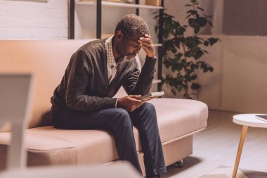 selective focus of depressed african american man sitting on sofa and holding photo frame clipart