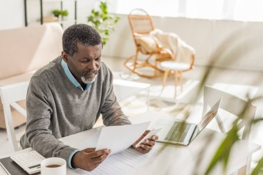 Üst düzey Afro-Amerikan bir adamın dizüstü bilgisayarın yanında otururken kamu hizmeti faturasına bakmasının seçici odağı.