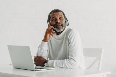 Kulaklıkla müzik dinleyen düşünceli Afro-Amerikan adam dizüstü bilgisayarın yanında oturmuş başka tarafa bakıyor.