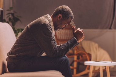 depressed african american man sitting on sofa at night and holding photo frame near head clipart