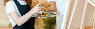 panoramic shot of kid holding palette and paintbrush near easel  clipart