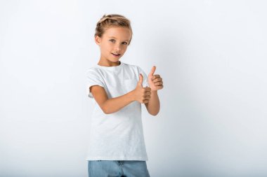 cheerful kid looking at camera and showing thumb up on white  clipart